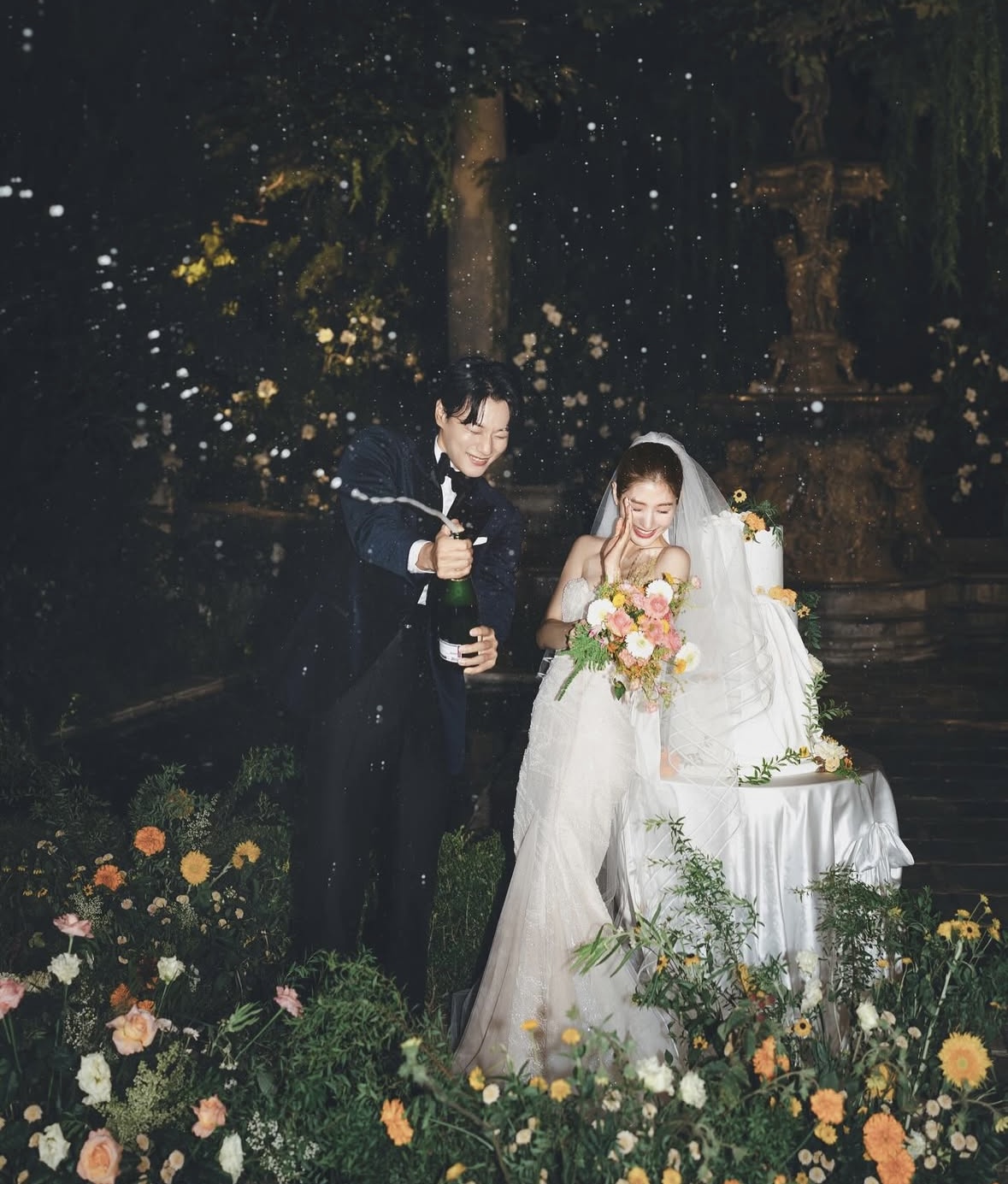 밤 정원 세트에서 신랑이 샴페인을 터뜨리고 베일 쓴 신부가 부케를 든 채 웃는 컷이에요.